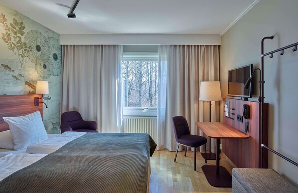 A hotel room with a bed, two chairs, a desk, a TV, and a window with curtains.