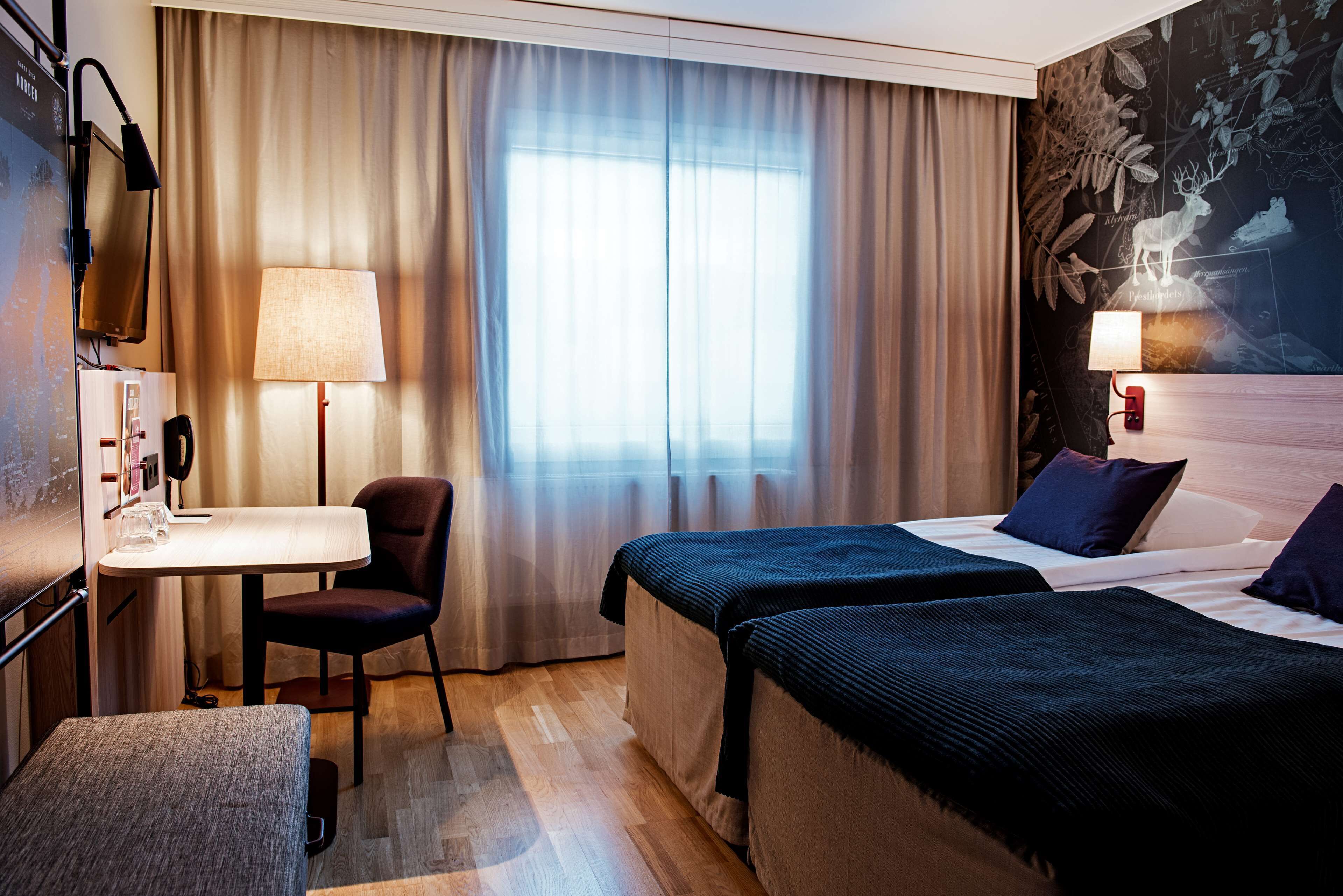 A hotel room with two beds, a desk with a chair, a lamp, and a large window with curtains.