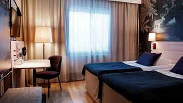A hotel room with two beds, a desk with a chair, a lamp, and a large window with curtains.