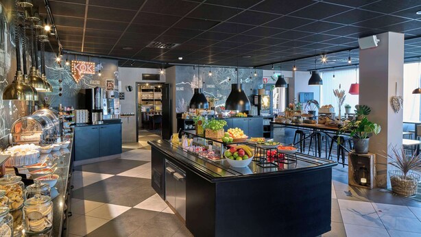 A modern cafe with a central island displaying fruits and pastries, a coffee machine, and a seating area with black chairs. - Scandic Södertälje (Sodertalje)