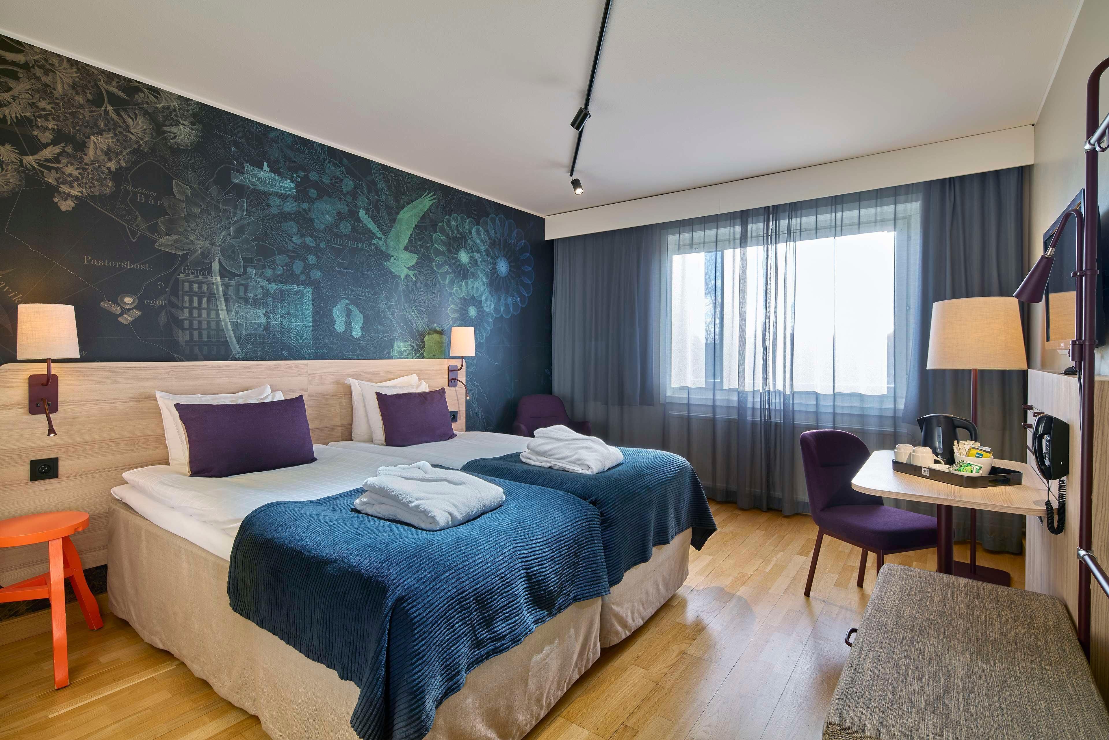 A hotel room with two beds, a desk with a telephone, a chair, and a large window with curtains.
