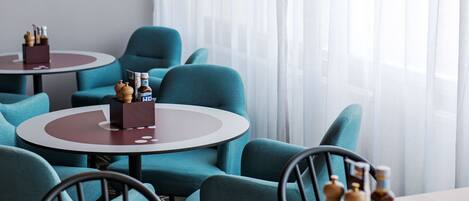 A modern interior with teal chairs and round tables, featuring a minimalist design and a view of the city through large windows.