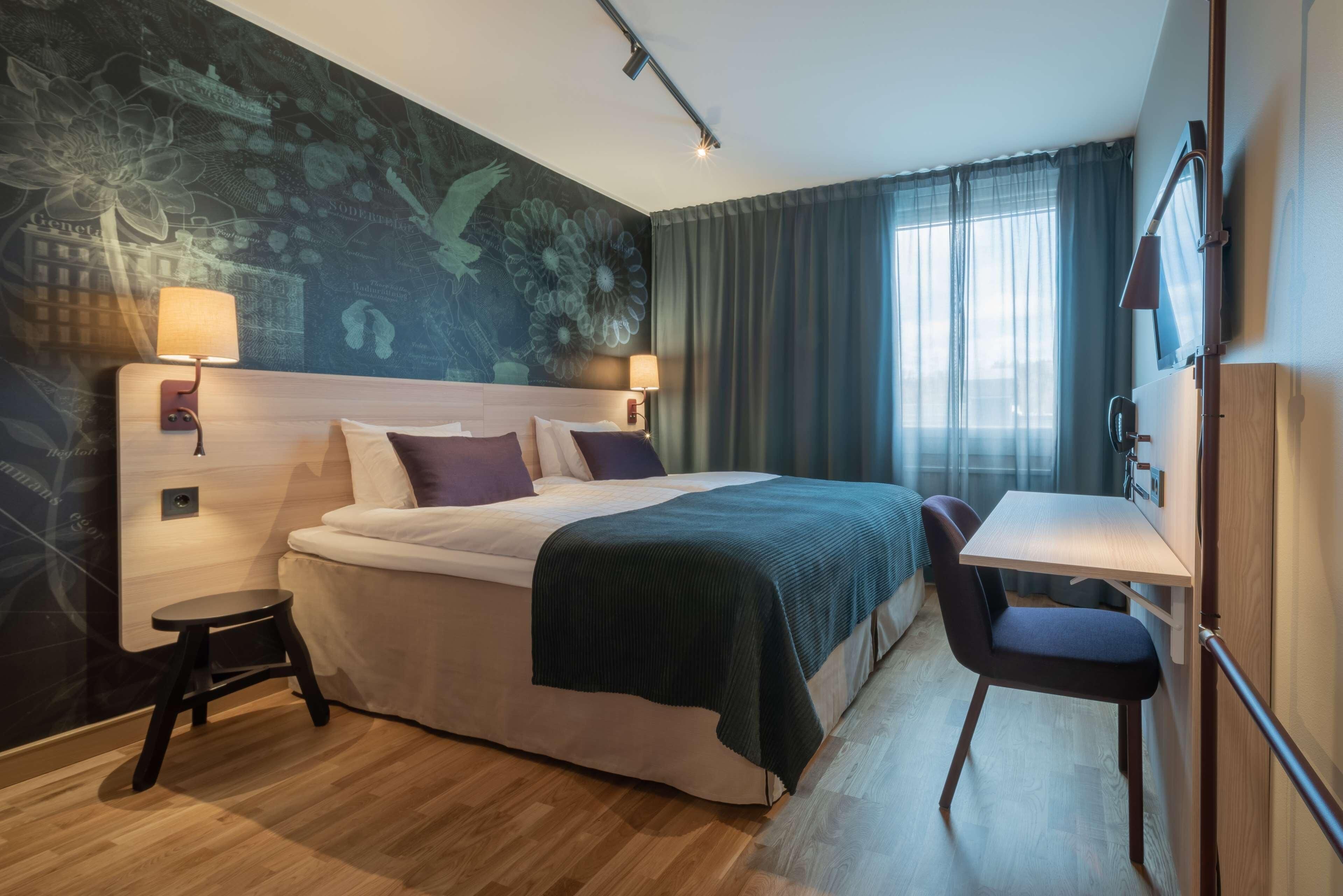 A hotel room with a bed, a desk, a chair, a lamp, a TV, and a window with curtains.