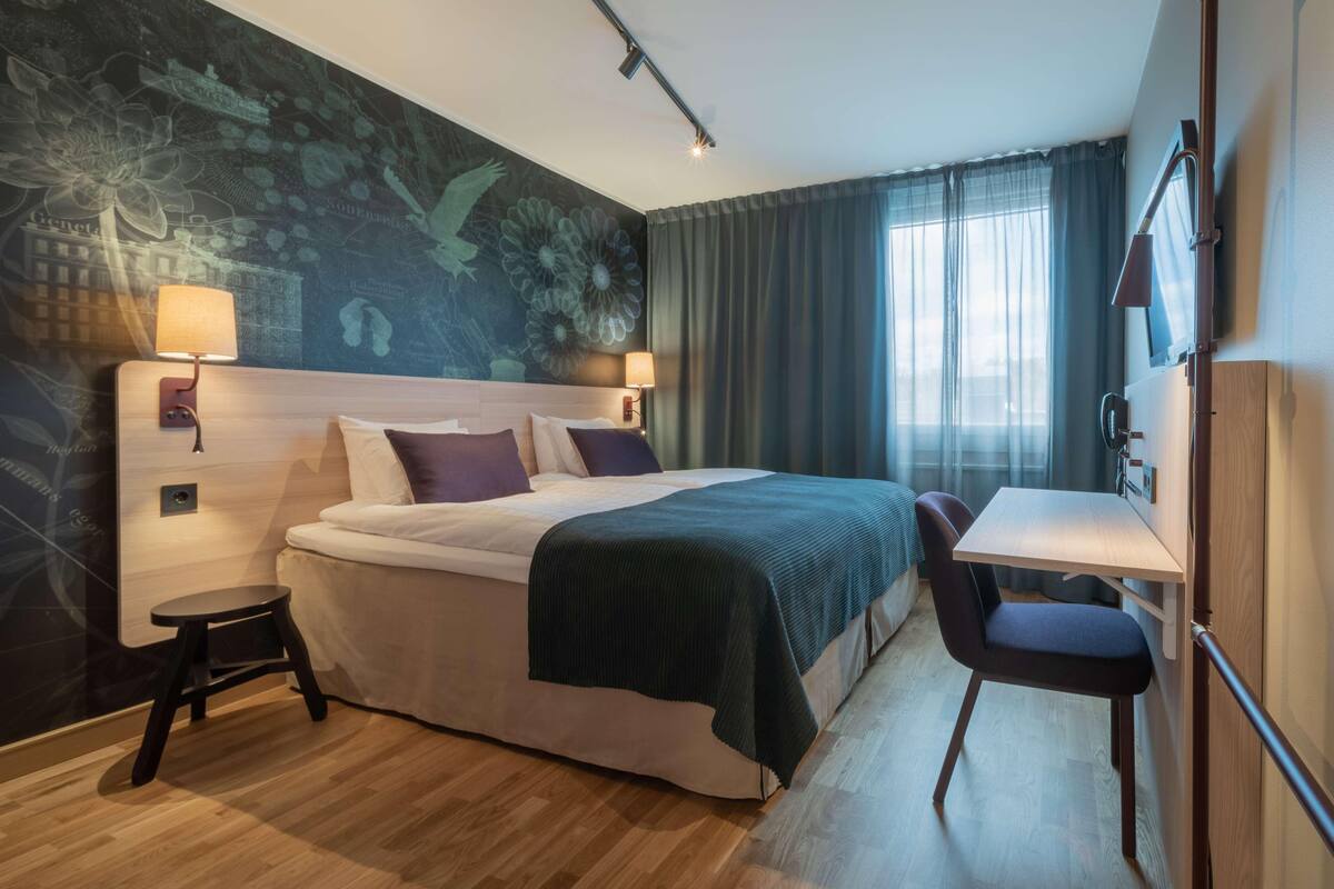 A hotel room with a bed, a desk, a chair, a lamp, a TV, and a window with curtains.