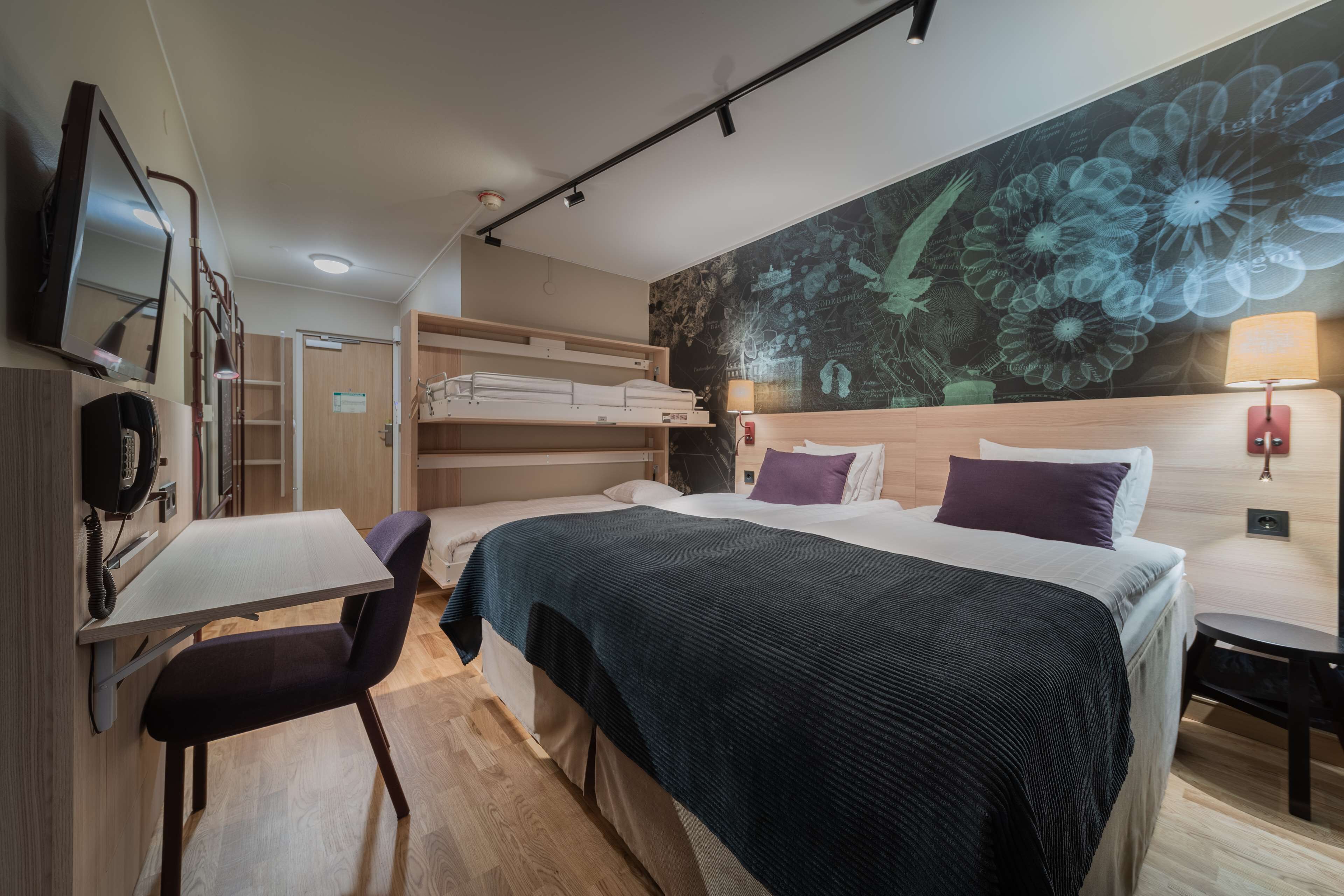 A modern hotel room with a bed, a desk, a chair, a TV, and a large floral mural on the wall.