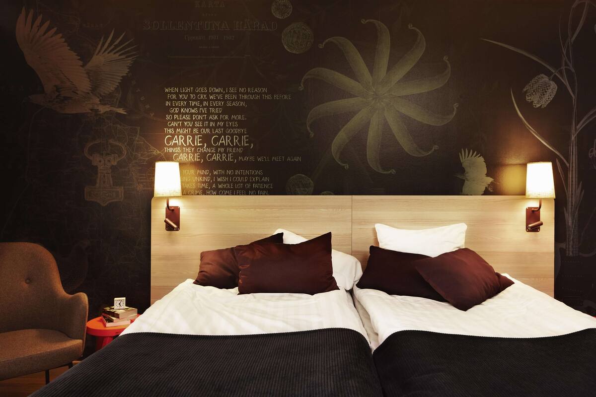 Et hotelværelse med sengegavl i træ, to senge med hvidt og brunt sengetøj, et lille bord med bøger og et ur, samt vægkunst med tekst og blomstermotiver.