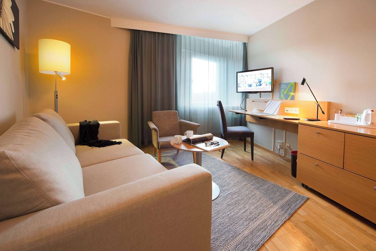 Et hotelværelse med en sofa, to stole, et skrivebord med en computer, en lampe og et fjernsyn.