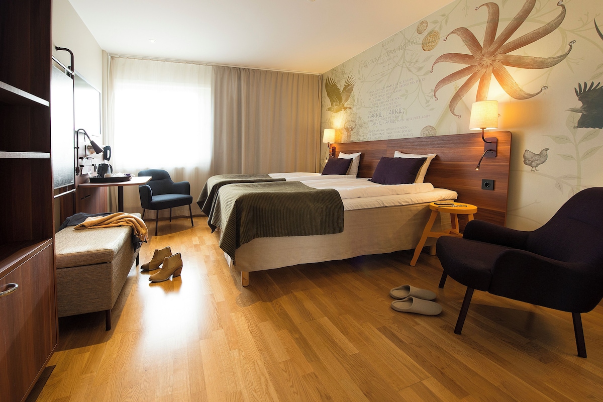 A hotel room with a bed, a desk, a chair, a small table, and a wall with a floral mural.