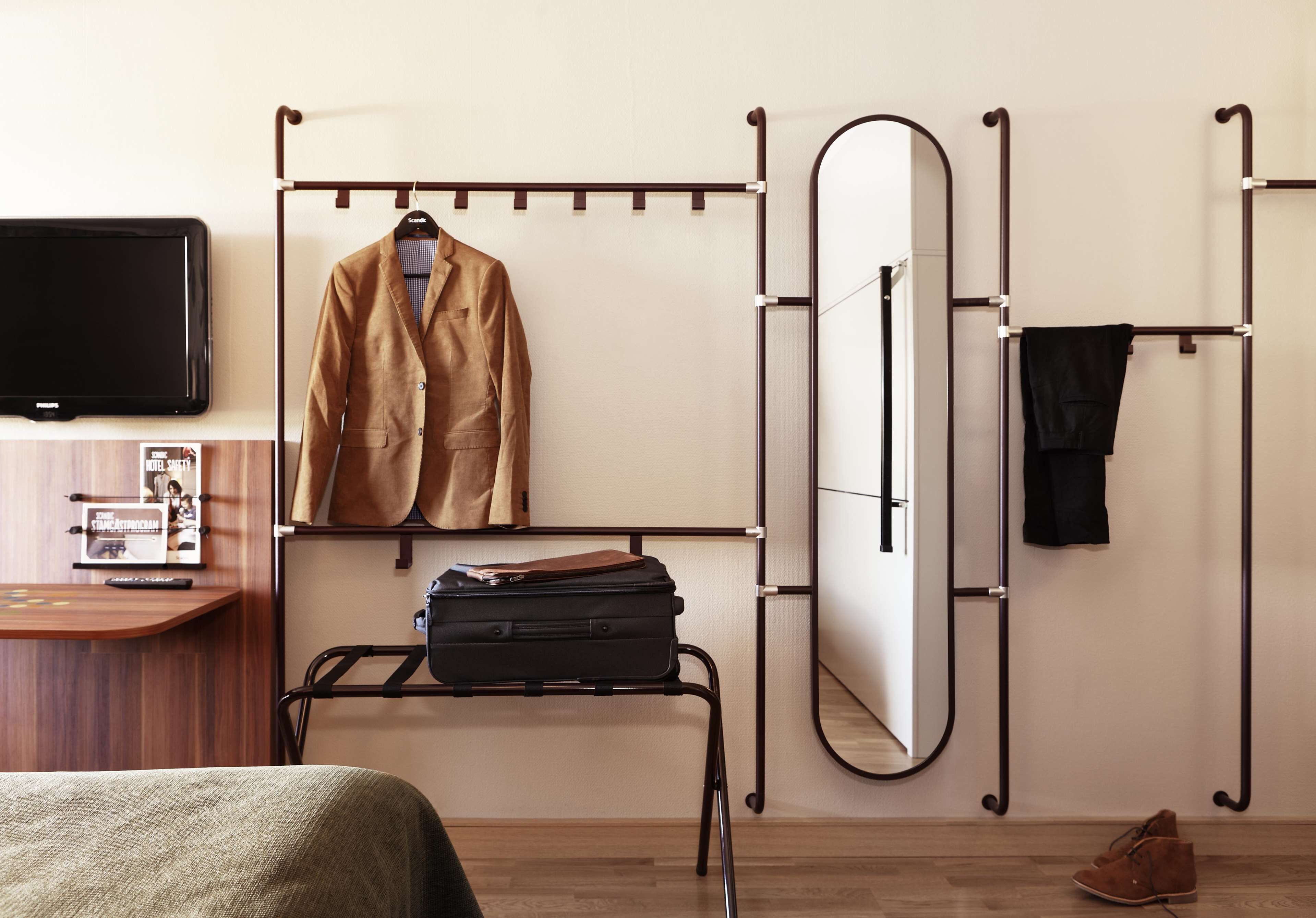 A hotel room with a bed, a desk with a flat-screen TV, a coat rack, a suitcase, a mirror, and a shoe rack.