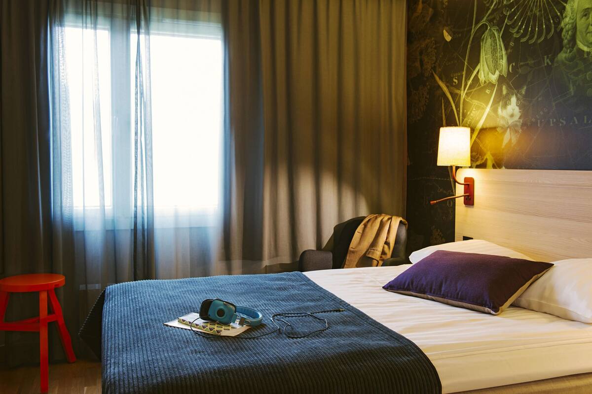A hotel room with a bed, a red stool, a lamp, and a wall with a map and plant.