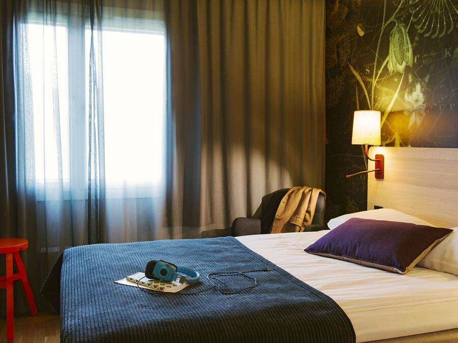 Quarto de hotel com uma cama, um banquinho vermelho, uma luminária e uma parede com um mapa e uma planta.