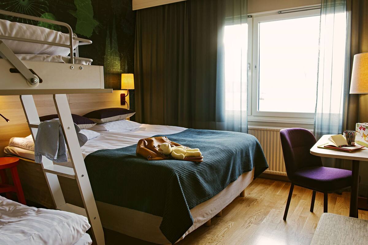 A hotel room with a bunk bed, a desk with a chair, a lamp, and a window with curtains.