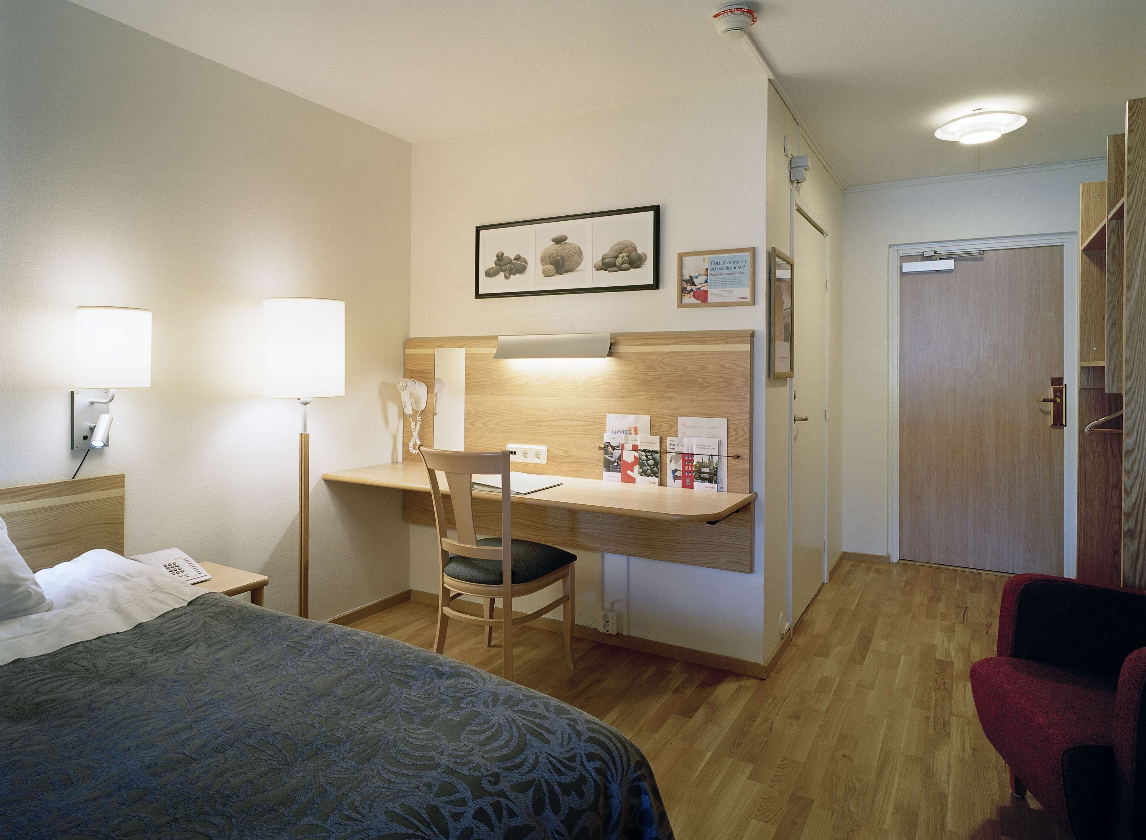 A hotel room with a bed, desk, chair, and a red armchair.