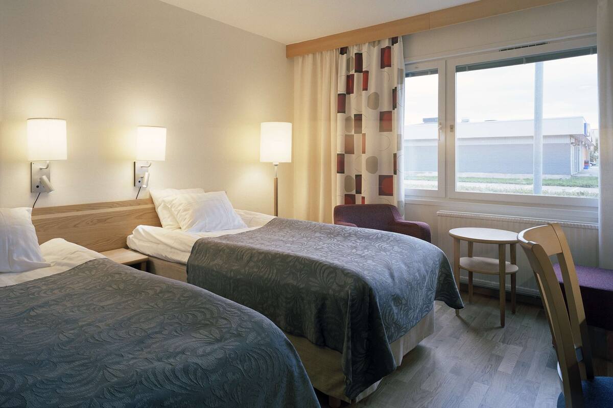 A hotel room with two beds, a small table, a chair, and a window with a view.