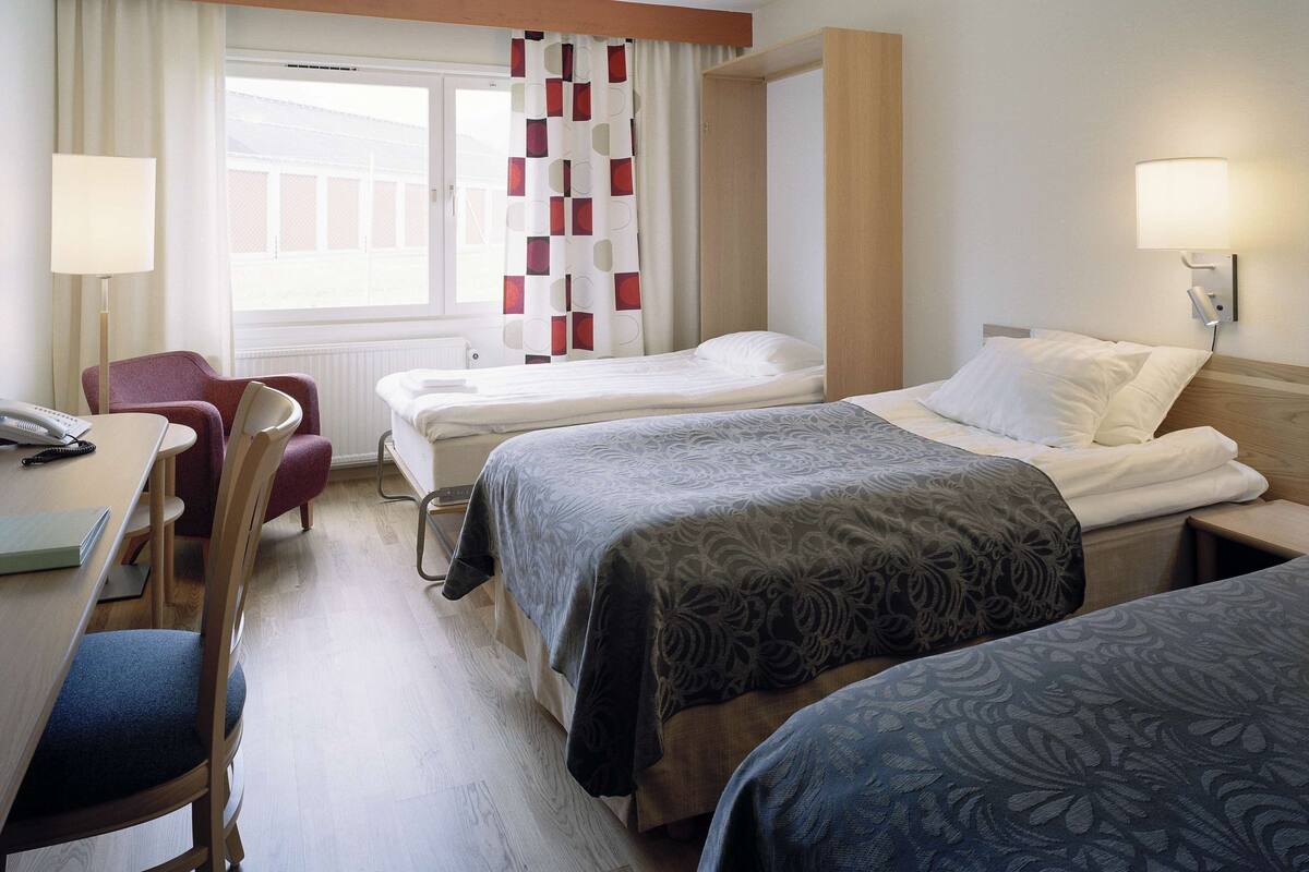 A hotel room with two single beds, a desk with a chair, a lamp, and a window with curtains.