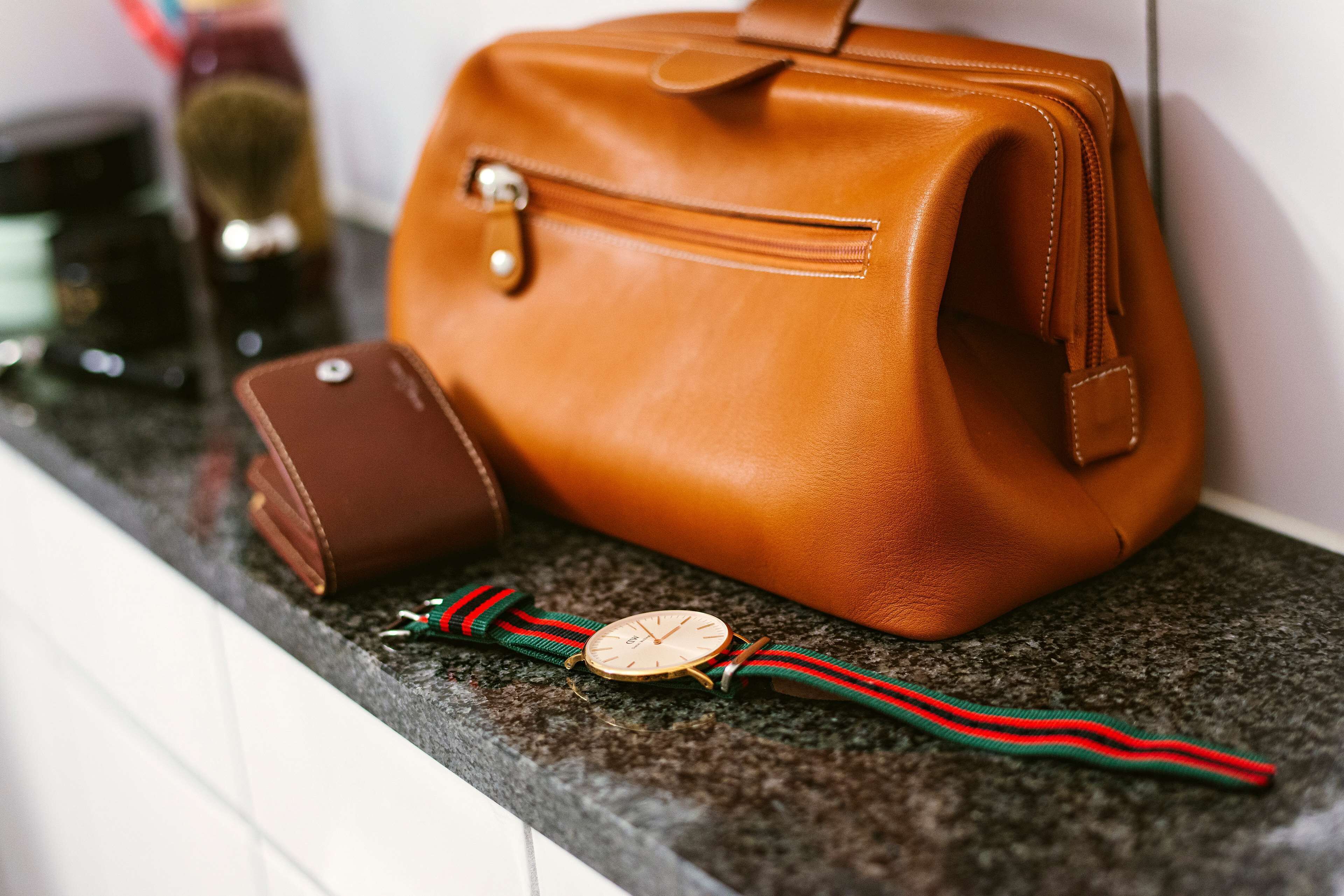 Un sac à main en cuir marron, un portefeuille assorti et une montre avec un bracelet vert et rouge, le tout posé sur un comptoir en granit.