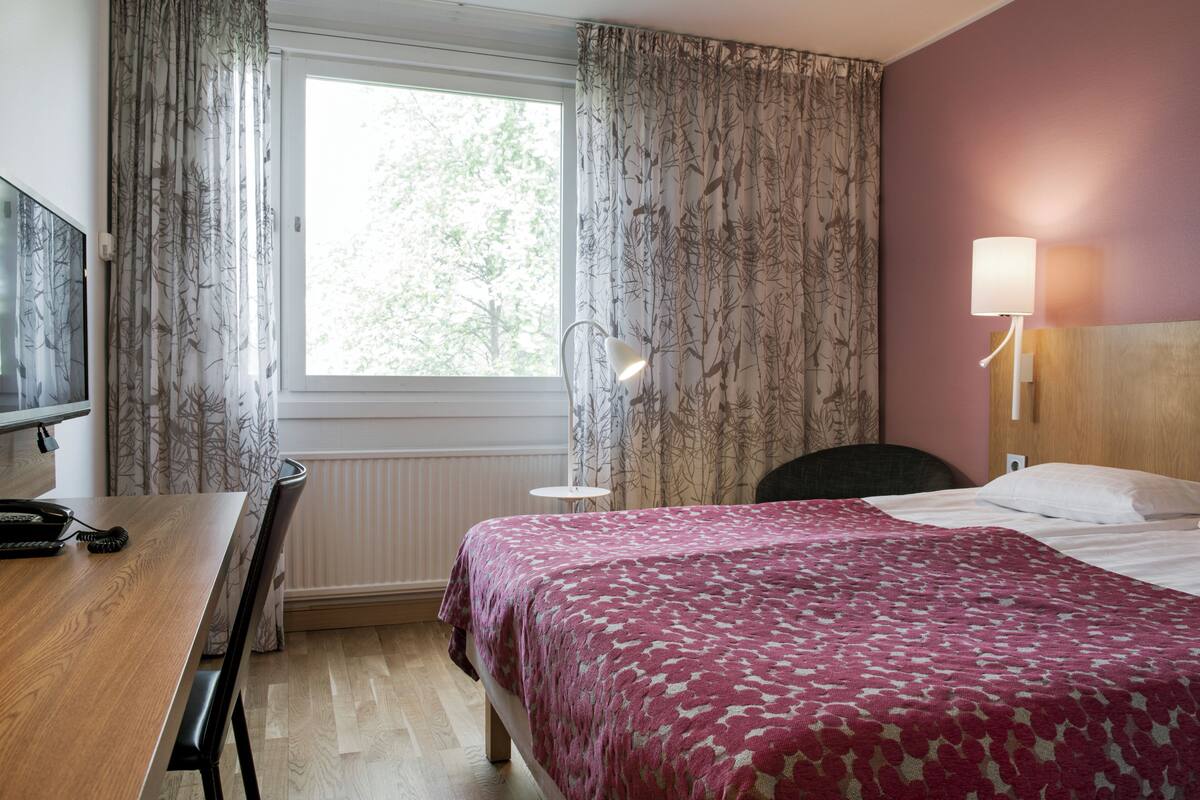A hotel room with a bed, a desk, a chair, a lamp, and a window with curtains.