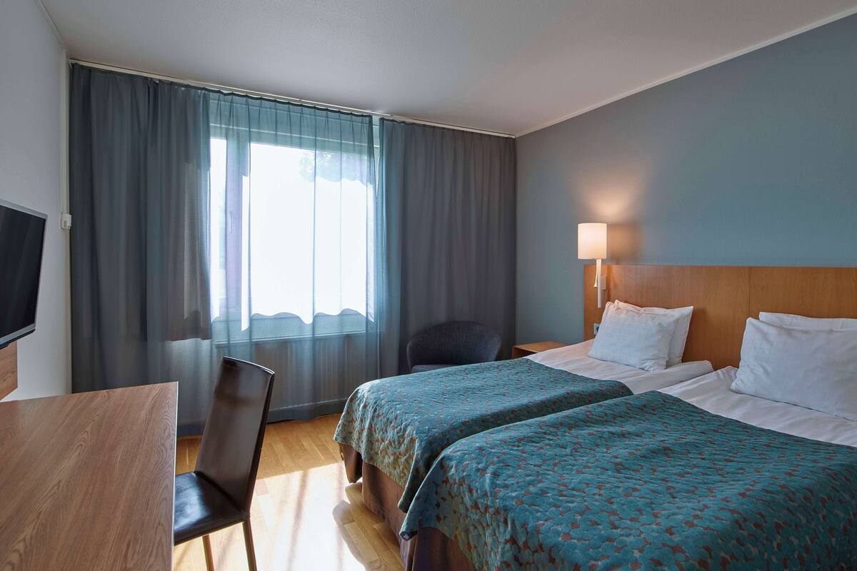 A hotel room with two beds, a desk, a chair, a TV, and a window with curtains.