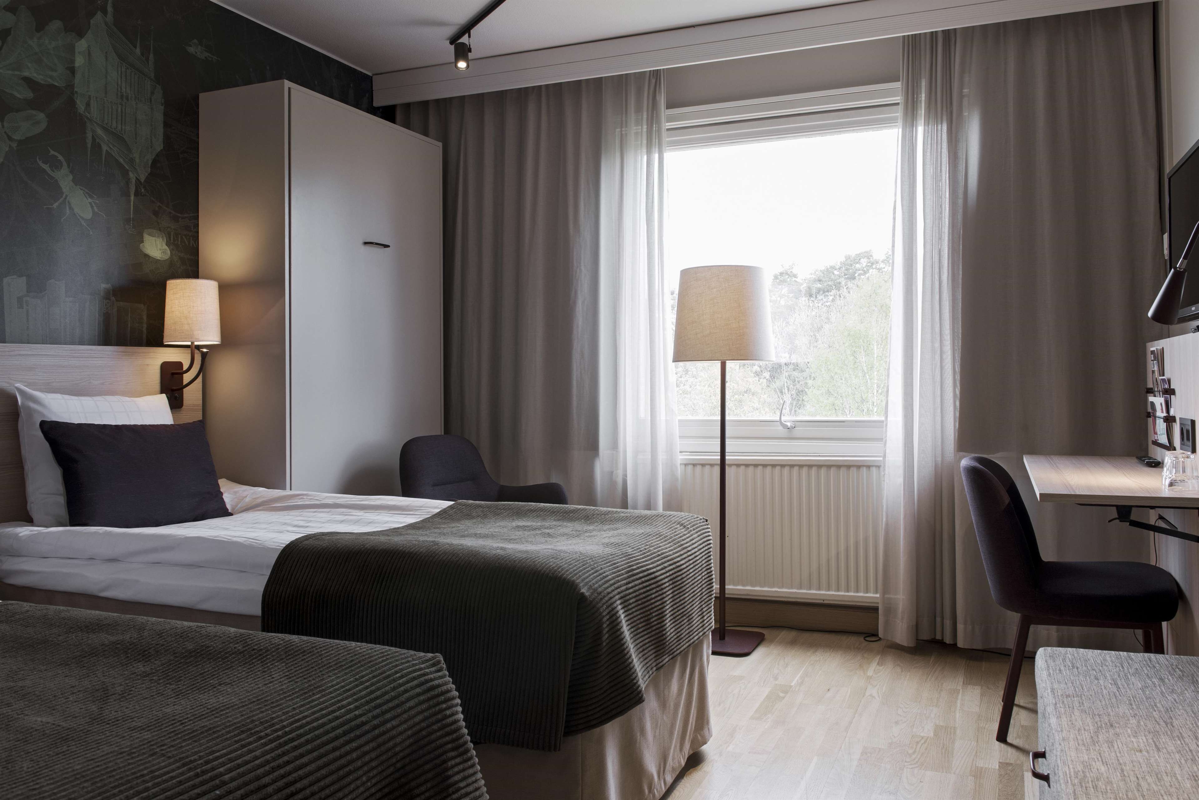 A hotel room with a bed, a desk, a chair, a lamp, and a view of the outdoors.