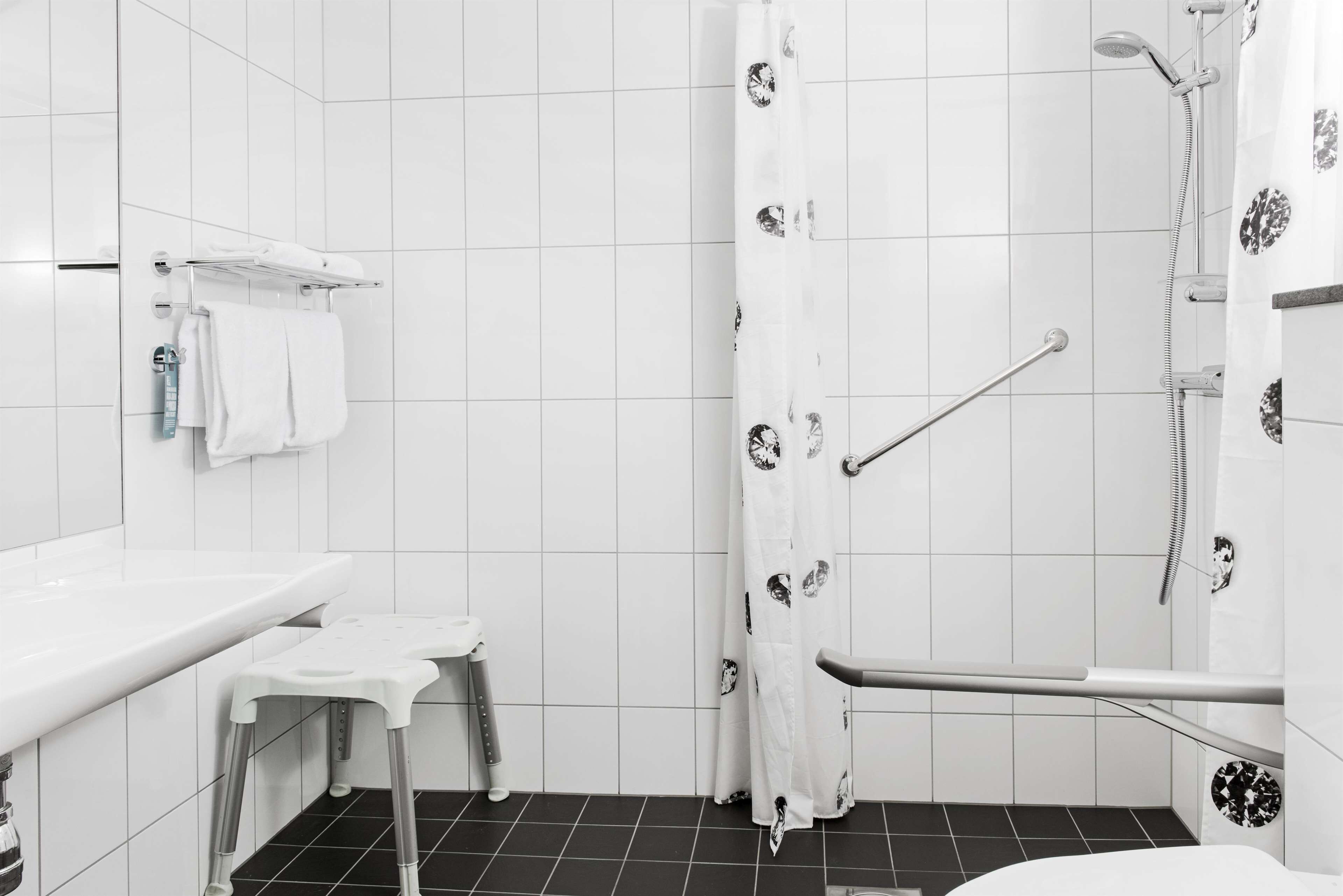 Un baño moderno con baldosas blancas, una ducha con cortina estampada, un lavamanos blanco y un taburete pequeño.