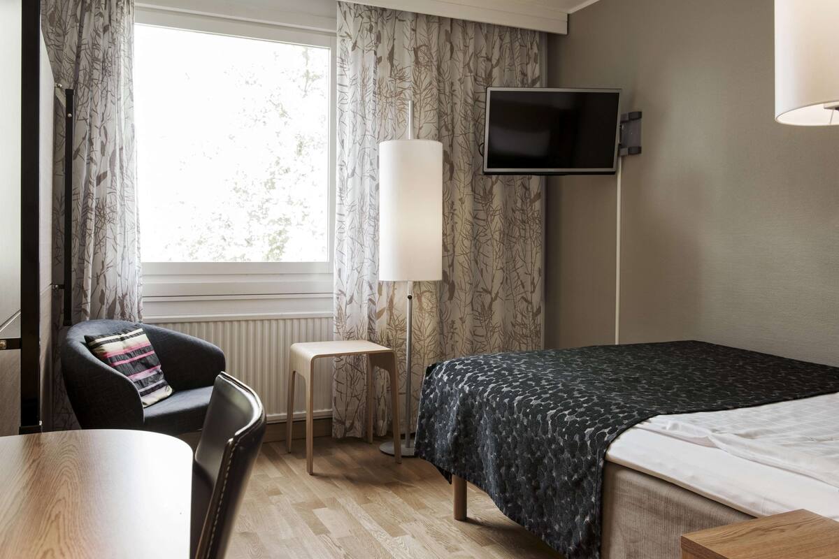 A hotel room with a bed, a chair, a small table, a TV mounted on the wall, and a window with curtains.