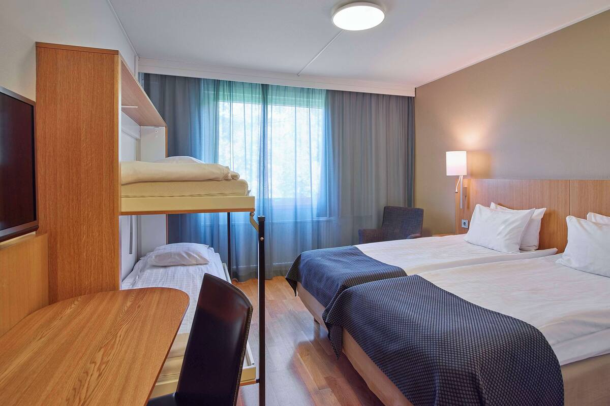 A hotel room with a bunk bed, a desk, a chair, a bedside table, a lamp, and a window with curtains.