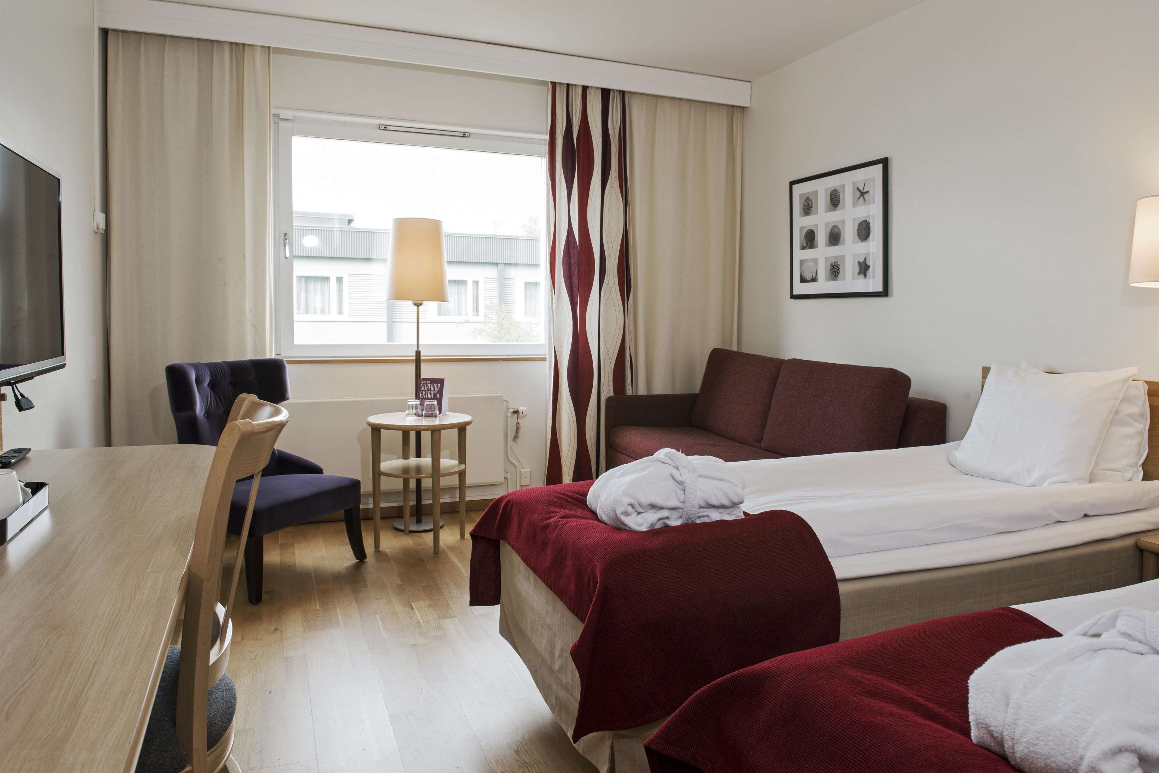 Quarto de hotel com uma cama, uma escrivaninha com cadeira, televisão, janela com cortinas e luminária.