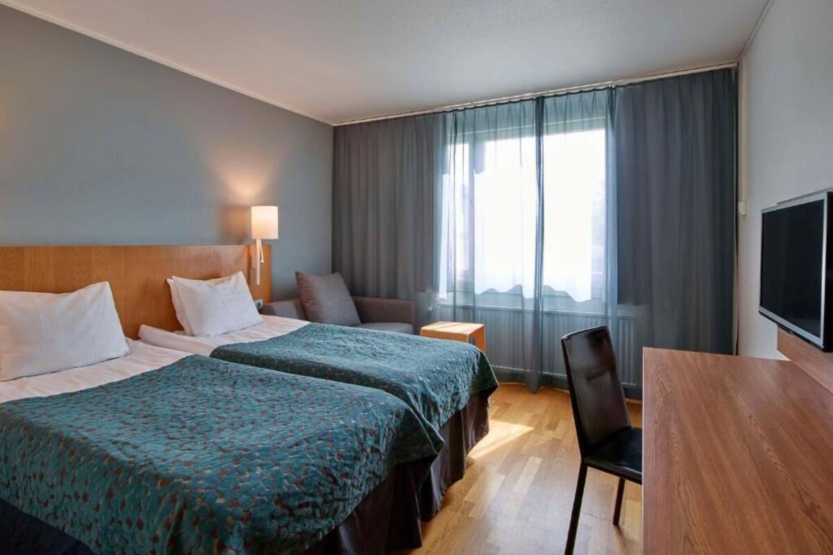 A hotel room with two beds, a desk, a chair, a TV, and a large window with curtains.