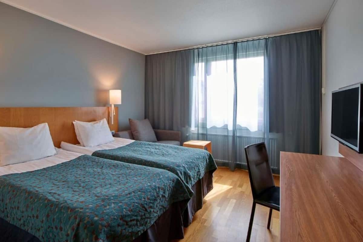 Habitación de hotel con dos camas, un escritorio, una silla, un televisor y un ventanal grande con cortinas.