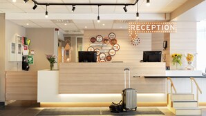 Un área de recepción con un mostrador de madera, un cartel que indica 'RECEPTION', una pared con varios iconos, un televisor y una escalera que conduce a un nivel superior.