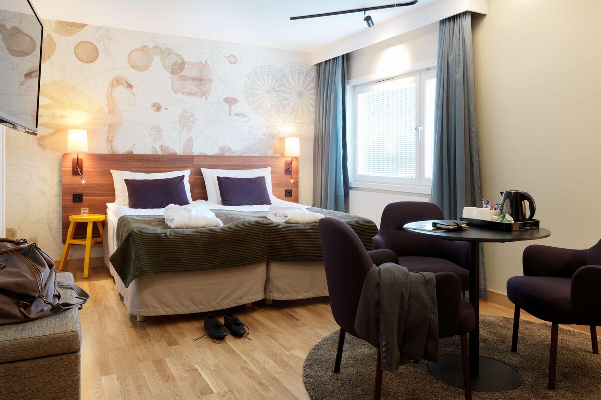 Una habitación de hotel con cama, dos sillas, una mesita, una lámpara y ventana con cortinas.