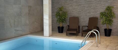 An indoor swimming pool with a tiled floor, two wicker chairs, and two potted plants.