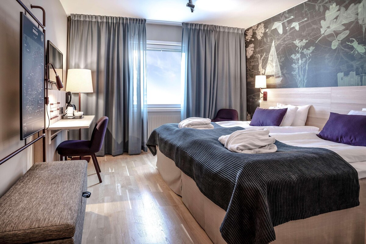 Hotellihuone, jossa on sänky, työpöytä, tuoli, penkki ja suuri ikkuna, jossa on verhot.