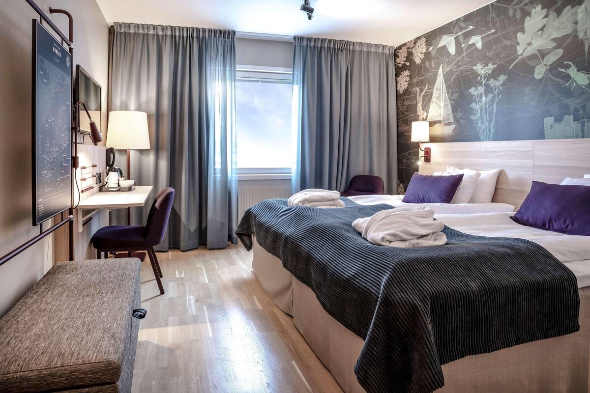 A hotel room with a bed, a desk, a chair, a bench, and a large window with curtains.