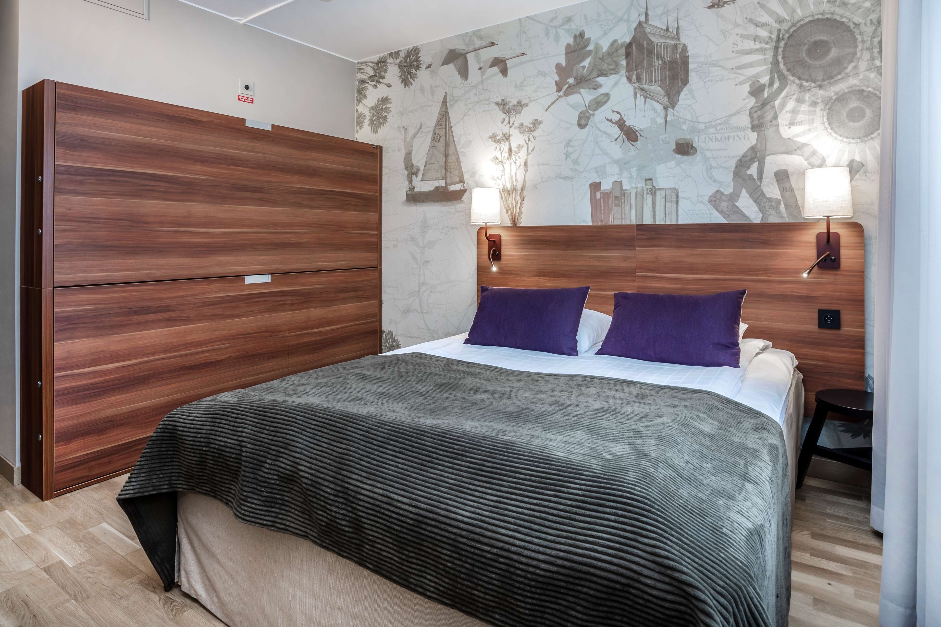 Un dormitorio con cabecera de madera, dos almohadas moradas, una colcha gris y un mural con motivos náuticos.