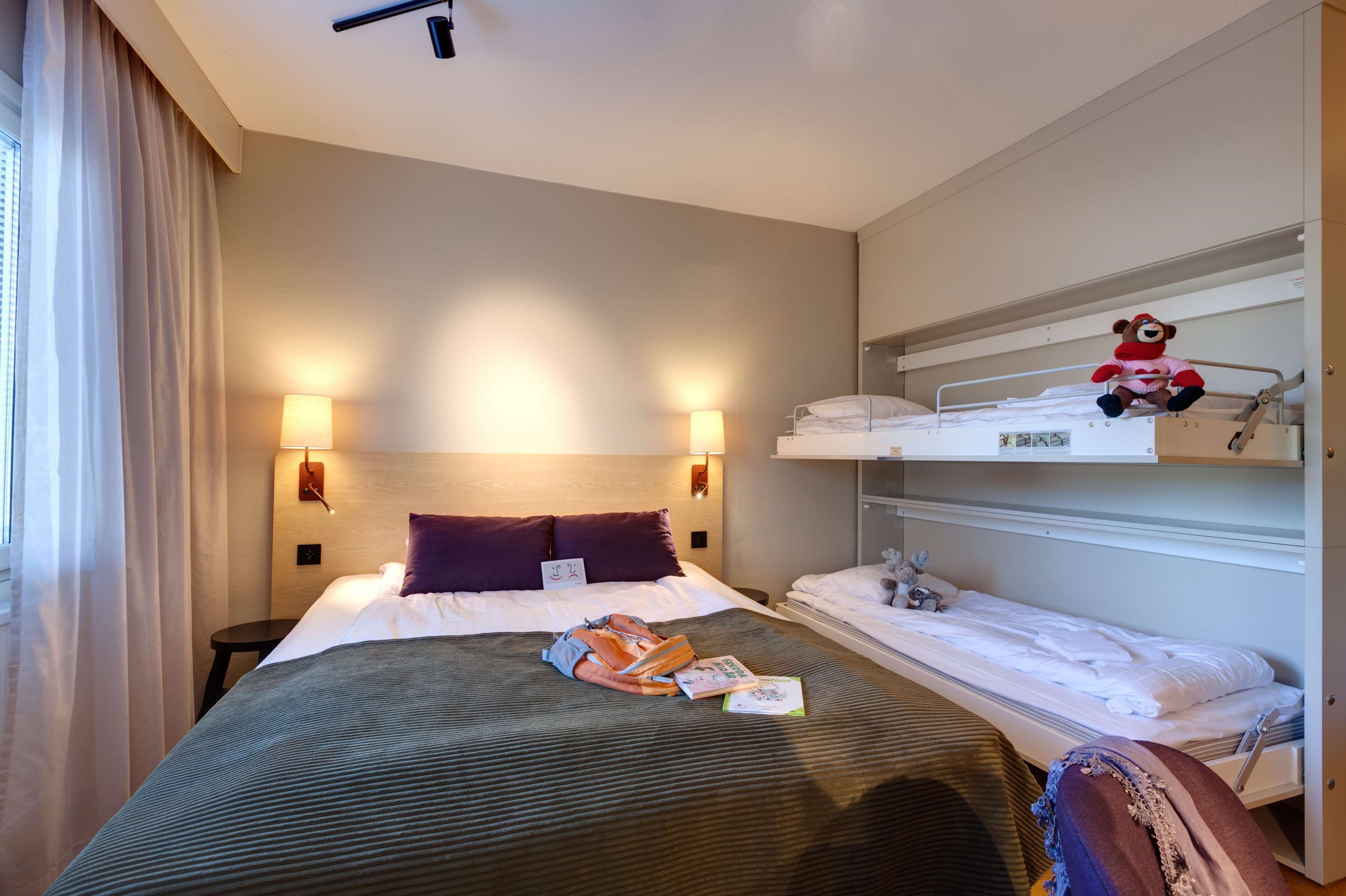 A hotel room with a large bed, two bedside tables, a bunk bed, and a window with curtains.