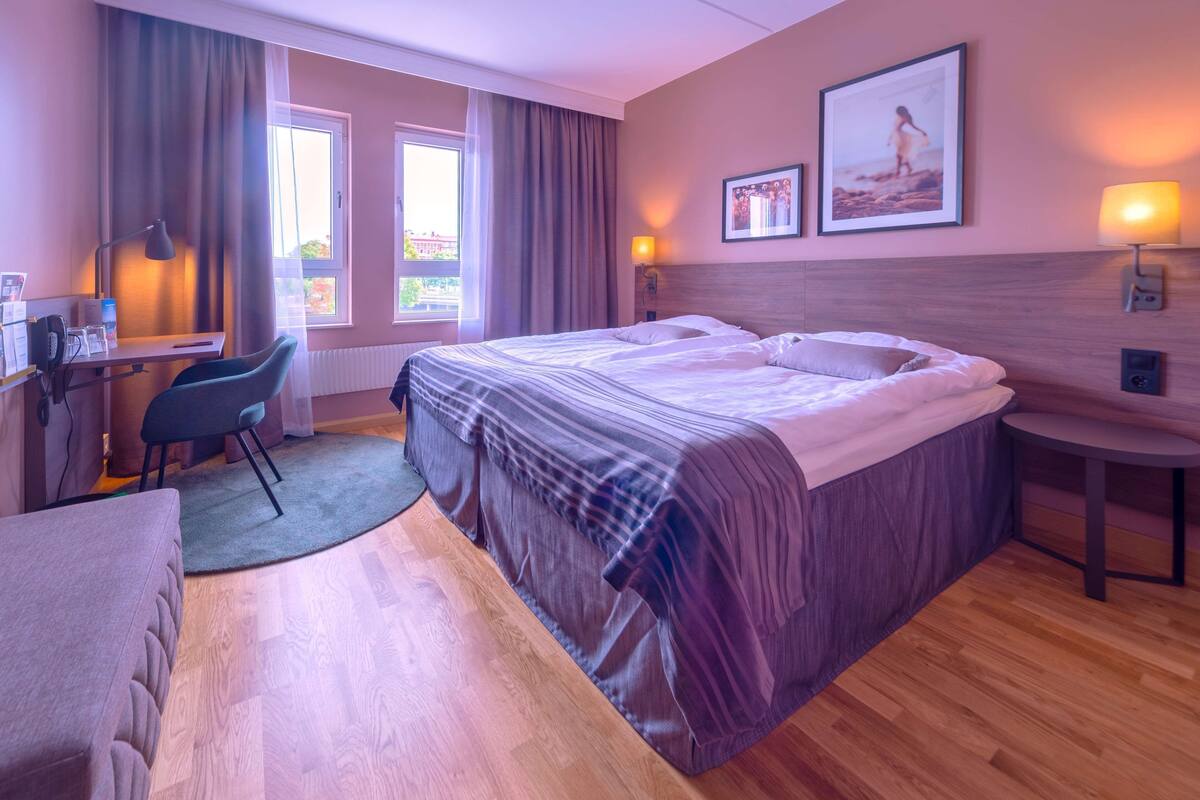 A hotel room with a bed, desk, chair, and two windows.