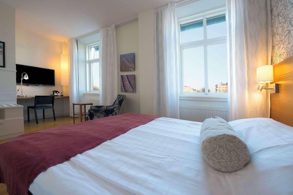 Une chambre d’hôtel avec un grand lit, un bureau, une chaise, une télévision et une fenêtre avec des rideaux.