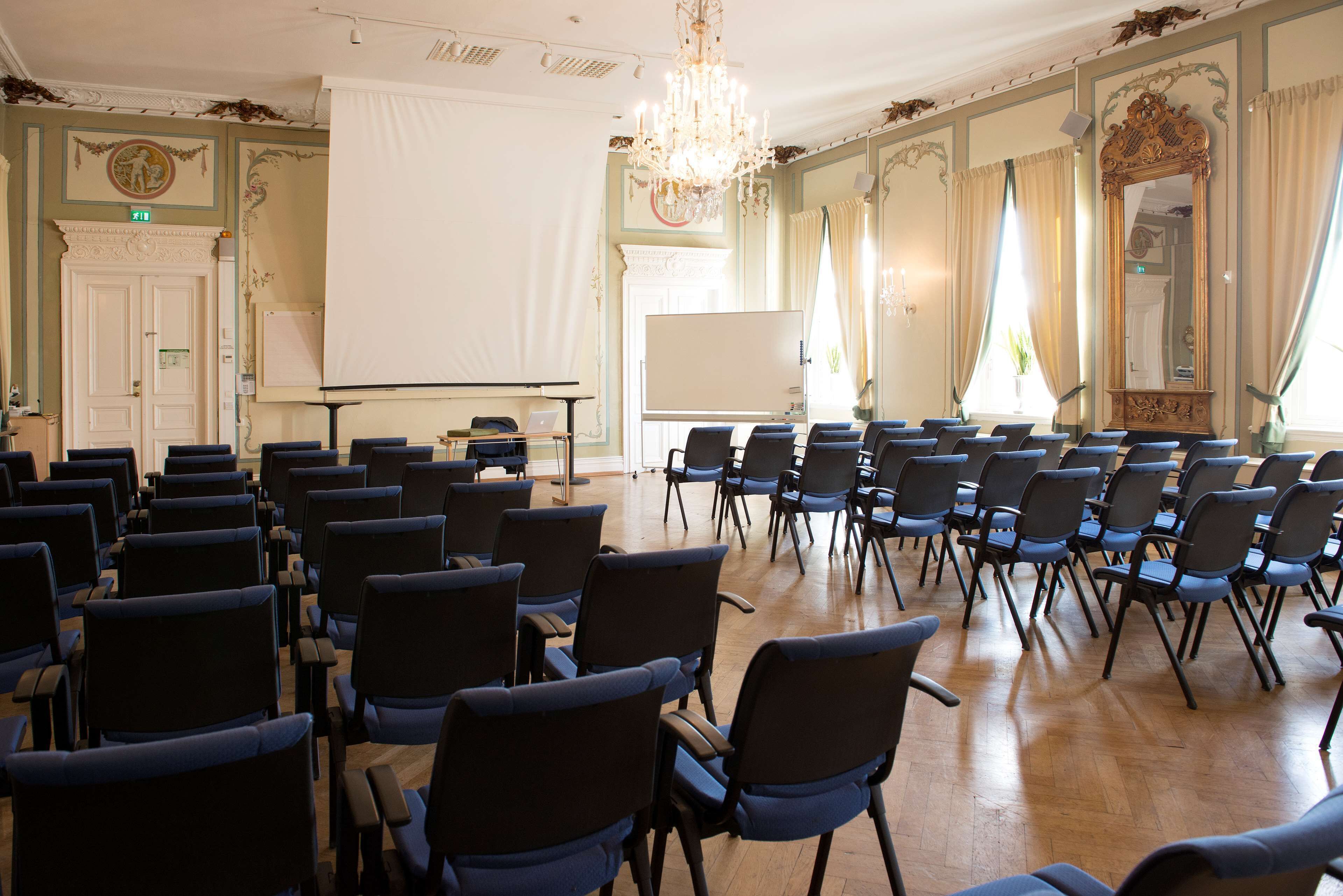 Une salle de conférence avec des rangées de chaises, un tableau blanc et un lustre.