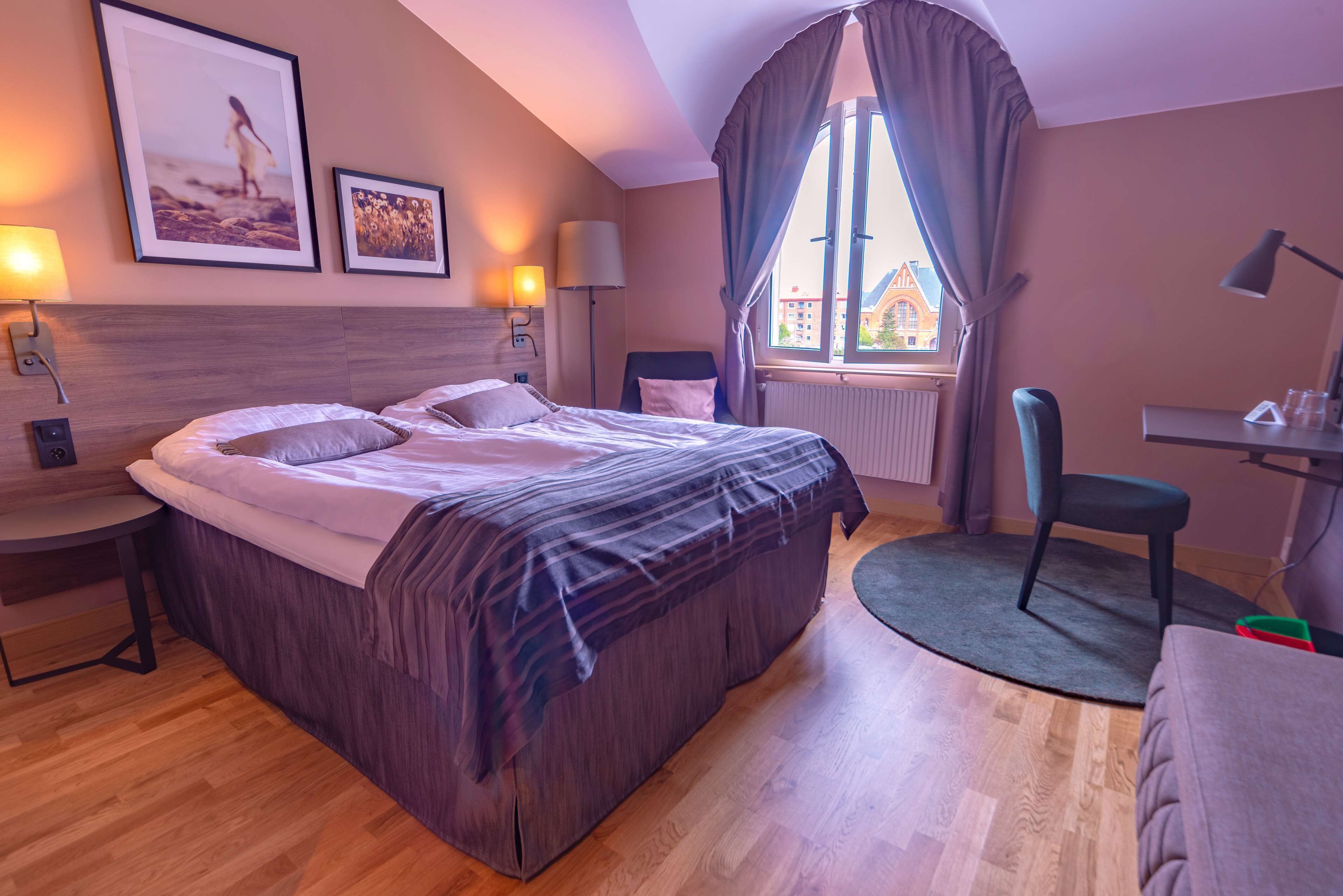 A hotel room with a bed, bedside tables, a desk, and a chair. There are two framed pictures on the wall, a window with curtains, and a view of buildings outside.