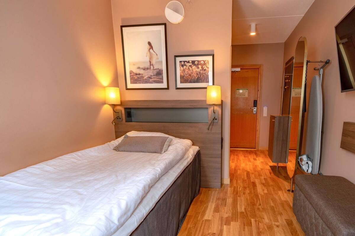 A hotel room with a bed, two wall-mounted lamps, a framed picture, a mirror, and a wooden door.