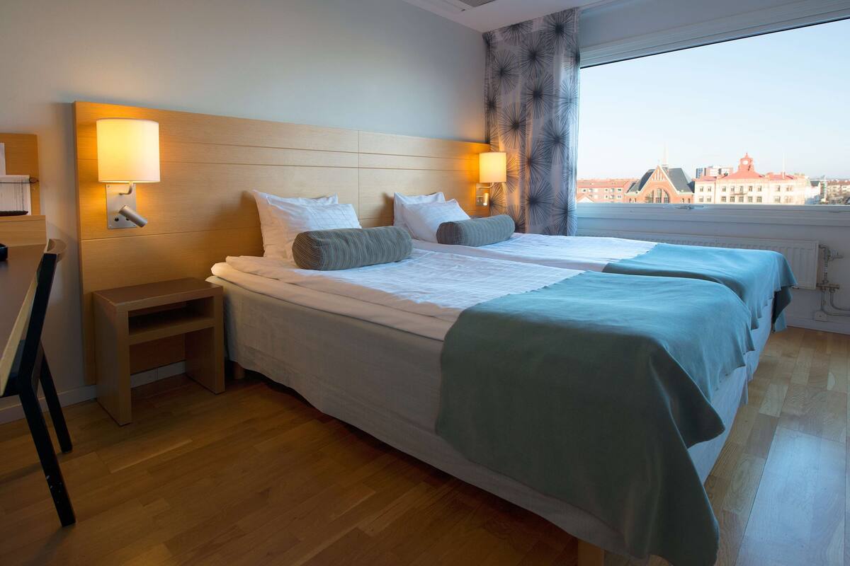 A hotel room with two beds, a wooden headboard, bedside lamps, a small wooden nightstand, and a view of buildings through a large window.