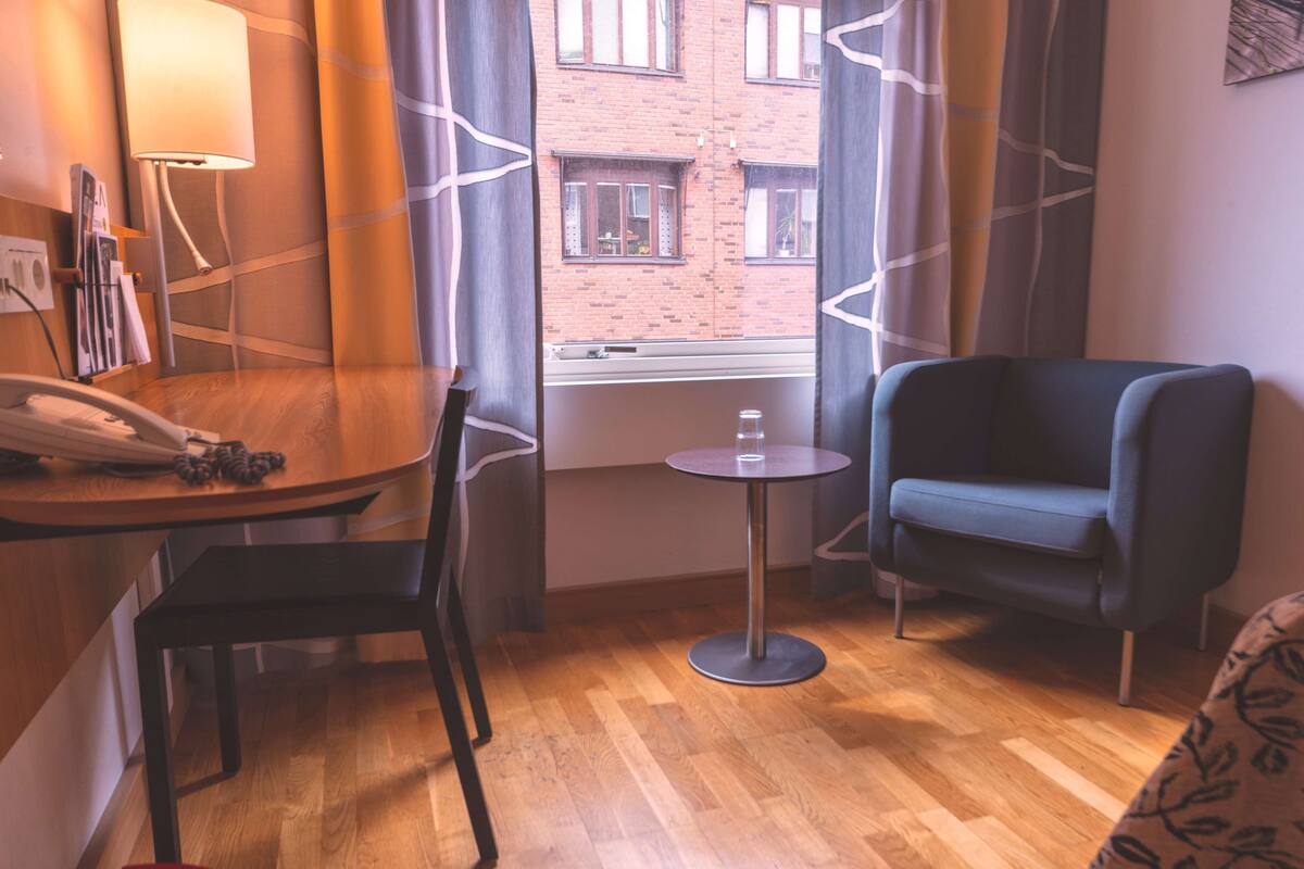 A hotel room with a wooden floor, a round table with a chair, a blue armchair, a small round table with a glass, and a window with a view of a brick building.