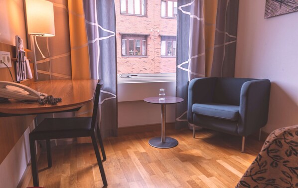 A hotel room with a wooden floor, a round table with a chair, a blue armchair, a small round table with a glass, and a window with a view of a brick building.