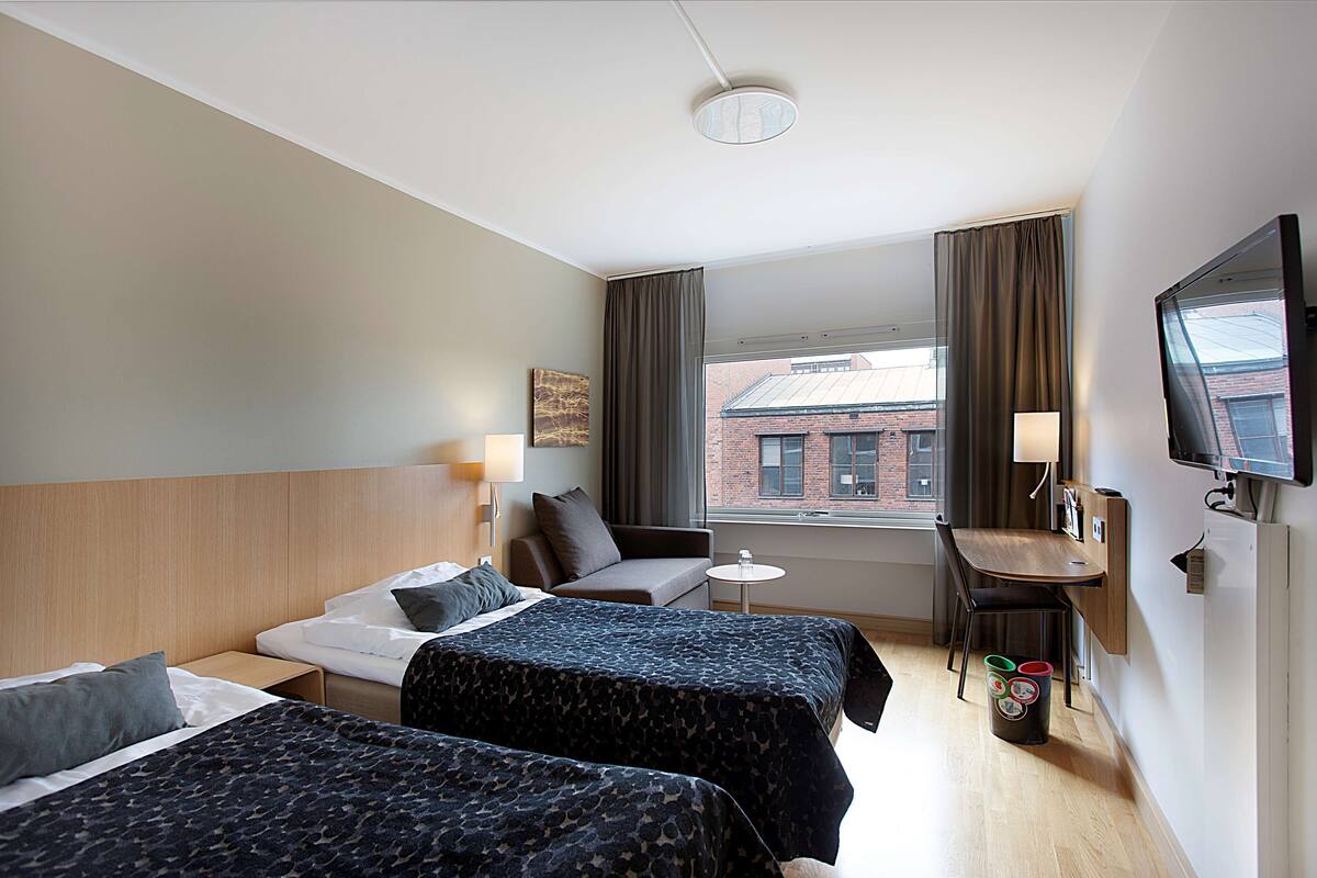 A hotel room with two beds, a desk, a chair, a TV, and a window with a view of a building.