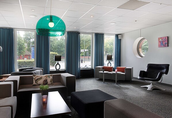 A modern hotel lobby with a sofa set, a round table, a black leather chair, and large windows with blue curtains.