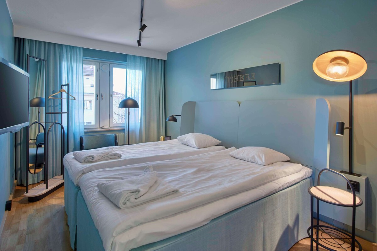 A hotel room with two beds, a TV, a desk, and a window with curtains.