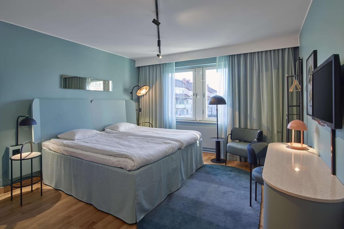 Una habitación de hotel moderna con una cama grande, un escritorio, una silla, un televisor y una ventana con cortinas.