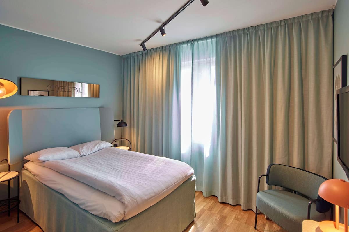 Un dormitorio con cama, una silla, un escritorio, una lámpara, un espejo y una ventana con cortinas.