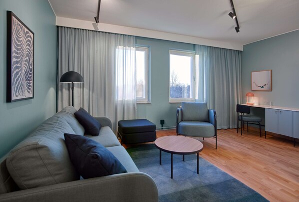 Una sala de estar moderna con un sofá, un reposapiés, una mesa de centro redonda y una silla. Hay dos ventanas con cortinas, un escritorio con una silla y una lámpara.